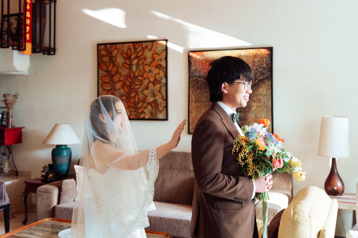 Bride in white veil adjusting grooms brown suit jacket holding flowers at indoor wedding ceremony.