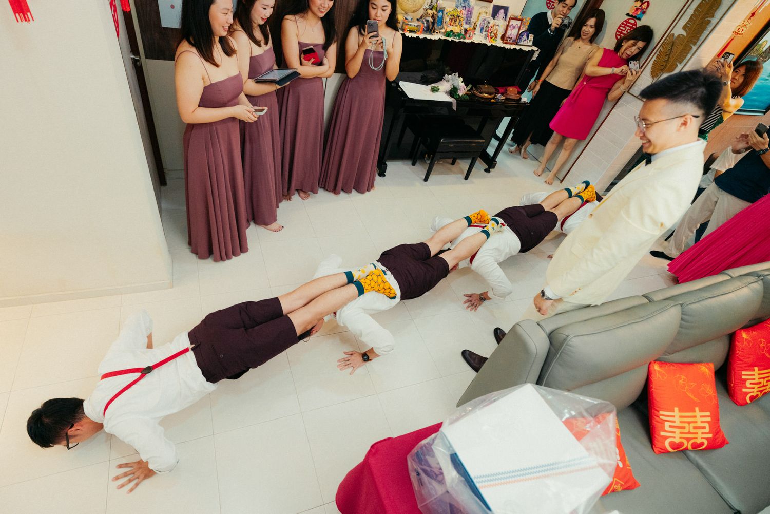 Men doing push-ups as a wedding game challenge while bridesmaids in mauve dresses watch and cheer indoors.