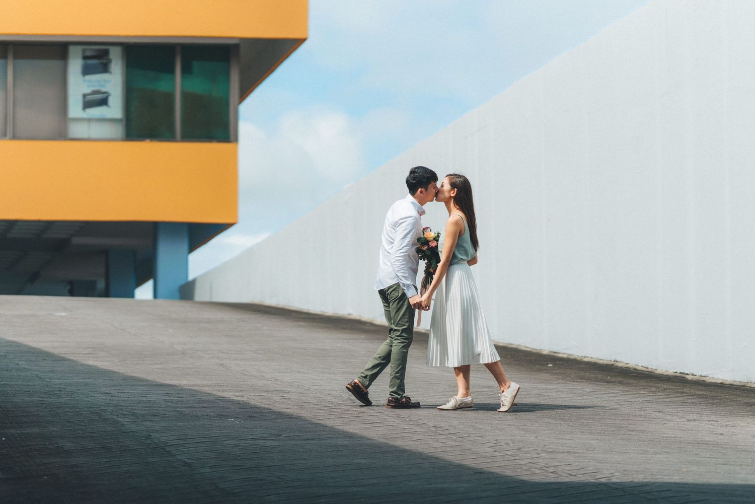 A couple in wedding attire kisses while standing on a sloped concrete surface against a bright sky.