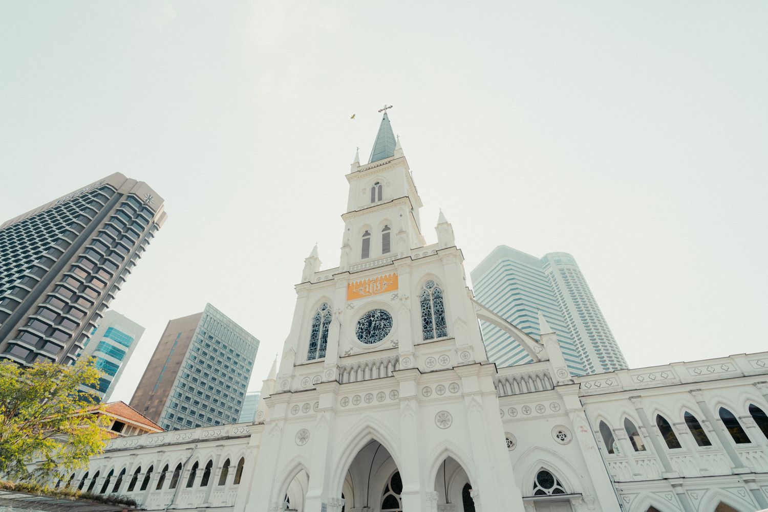 Historic white clock tower church with Gothic arches stands amid modern Singapore skyscrapers under a bright sky.