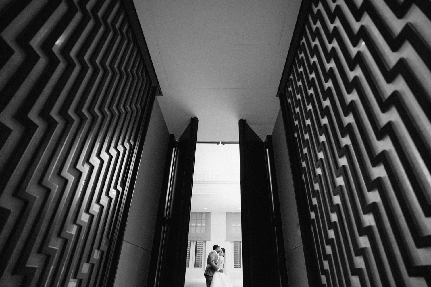 Black and white photo of a couple embracing in a doorway framed by dramatic zigzag patterned walls.