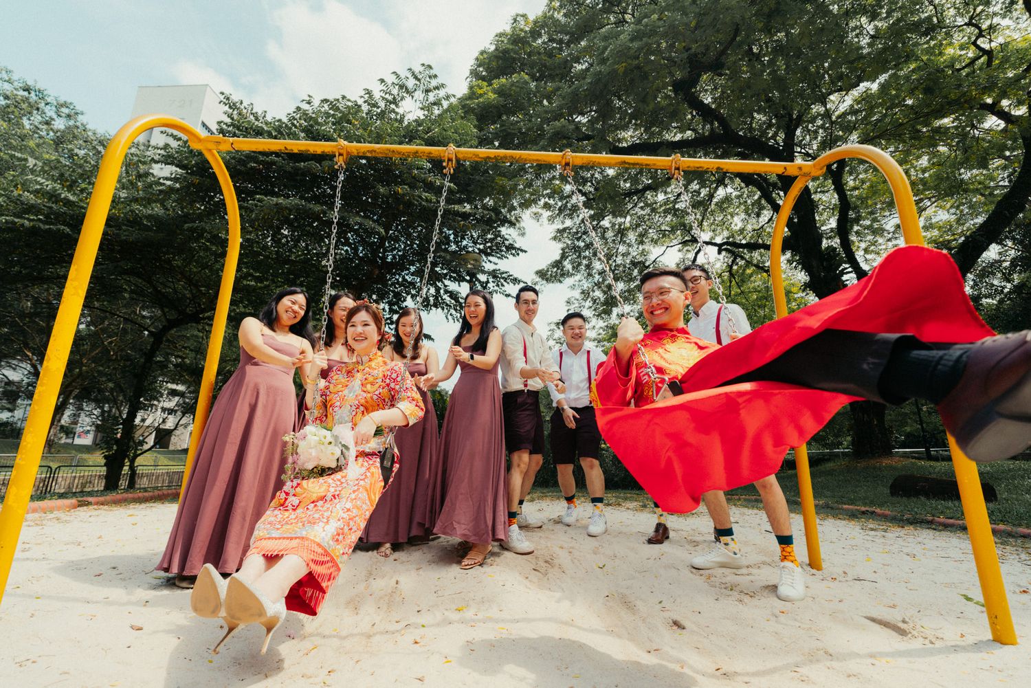 Wedding party posing playfully on yellow playground swings, bride in red qipao with flowing fabric, bridesmaids in mauve dresses.