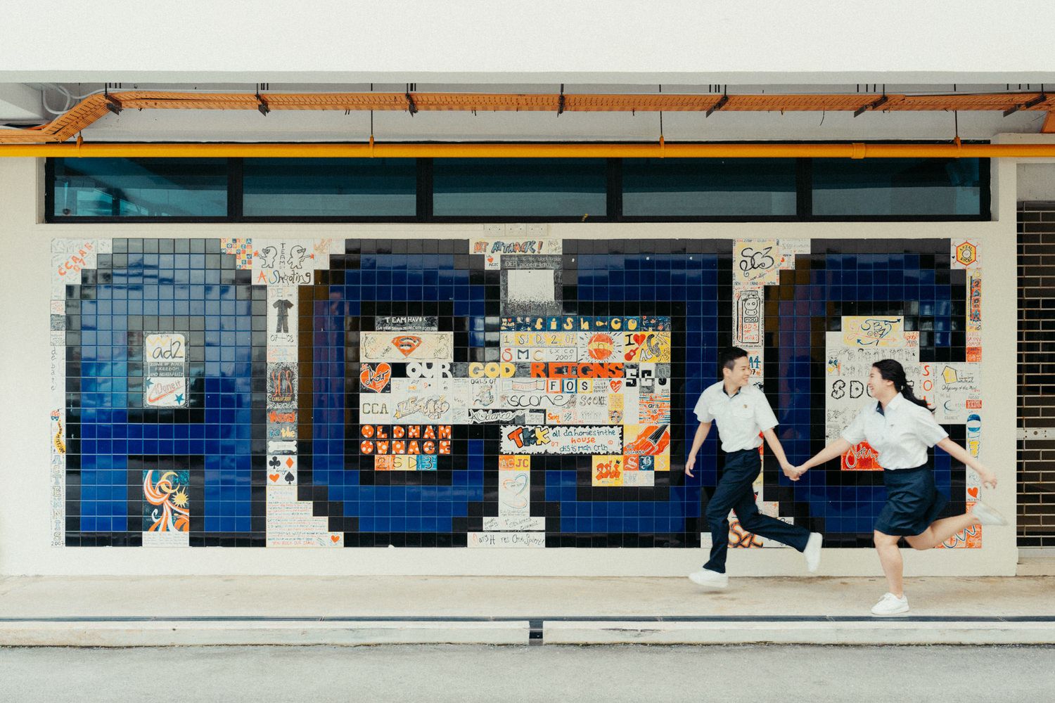Couple holding hands walking in front of a colorful mosaic tile mural spelling ACDC on a urban wall.