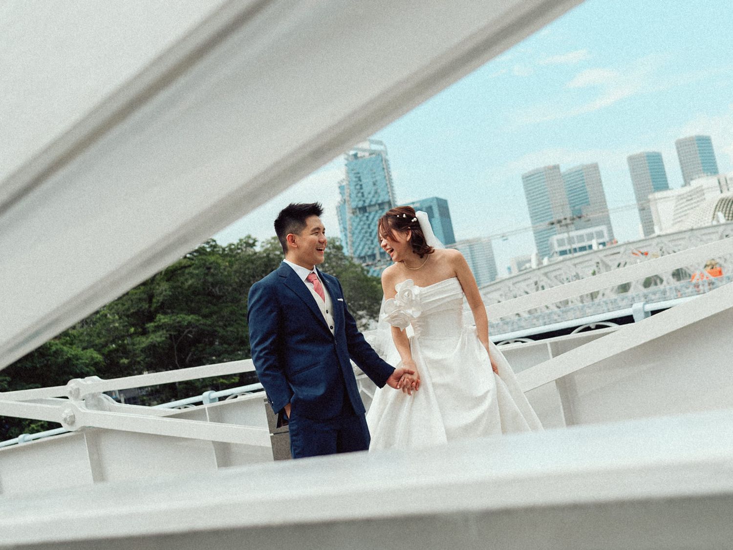 A couple in wedding attire share a moment on a white deck overlooking a city skyline.