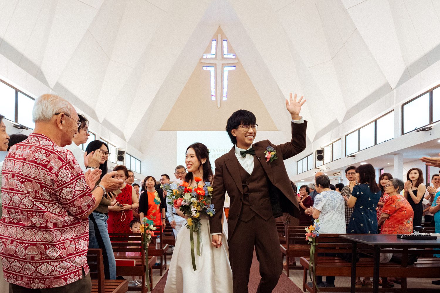 Newlywed couple walks down church aisle, groom waving, bride holding colorful bouquet, guests applauding in decorated chapel.