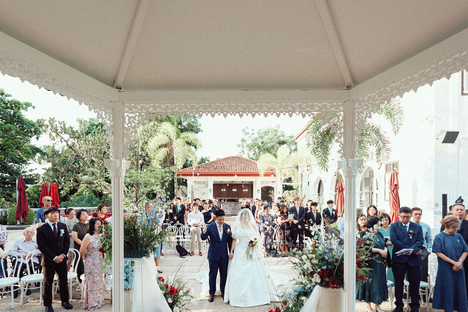 Outdoor wedding ceremony under an elegant white gazebo with palm trees and guests gathered in a tropical garden setting.