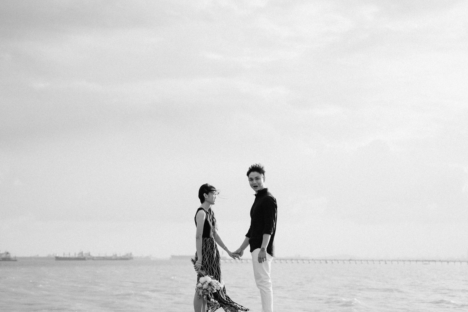 A couple stands together on a beach with a misty ocean view in black and white.