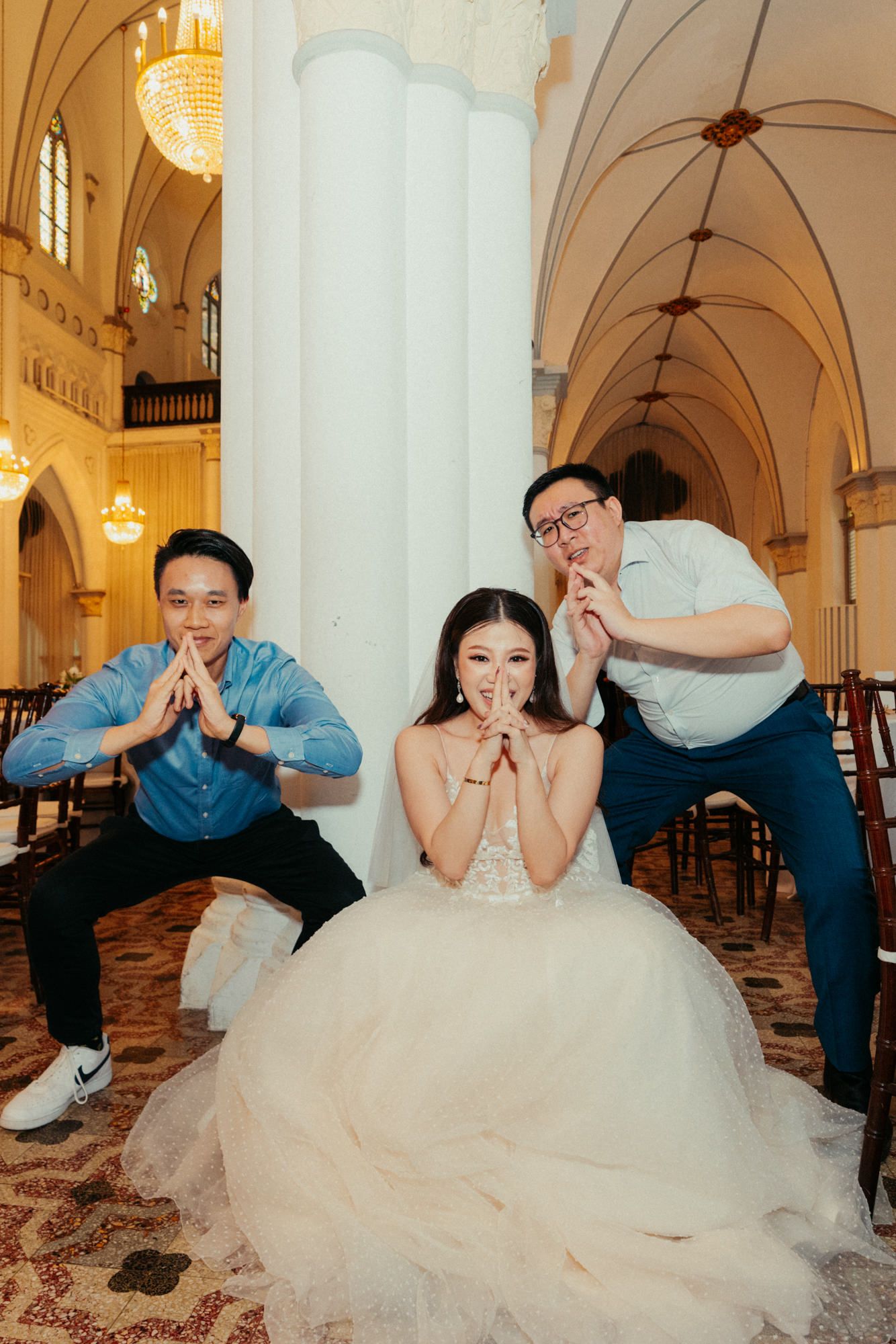 Bride in white gown poses playfully with two men inside an elegant church with arched ceilings and chandeliers.