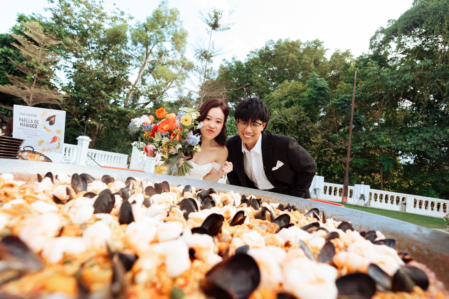 A bride and groom lean over a large seafood platter outdoors, surrounded by lush greenery on their wedding day.