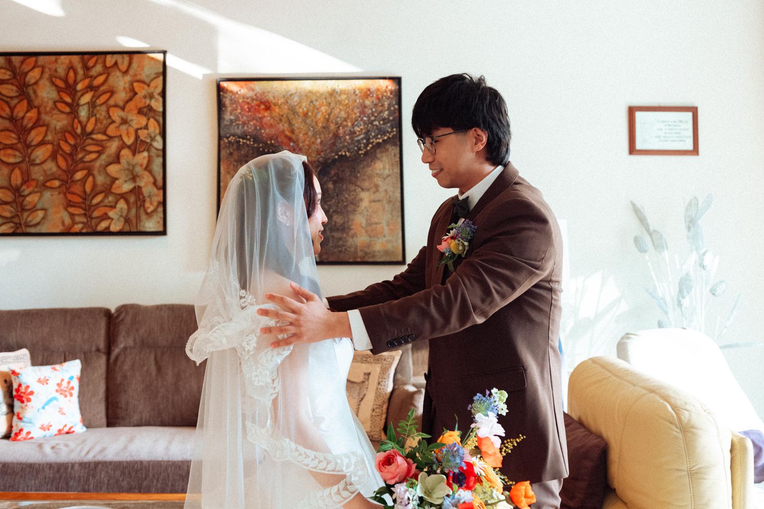 A groom in a brown suit adjusts the veil of his bride in a white dress during an intimate indoor wedding moment.