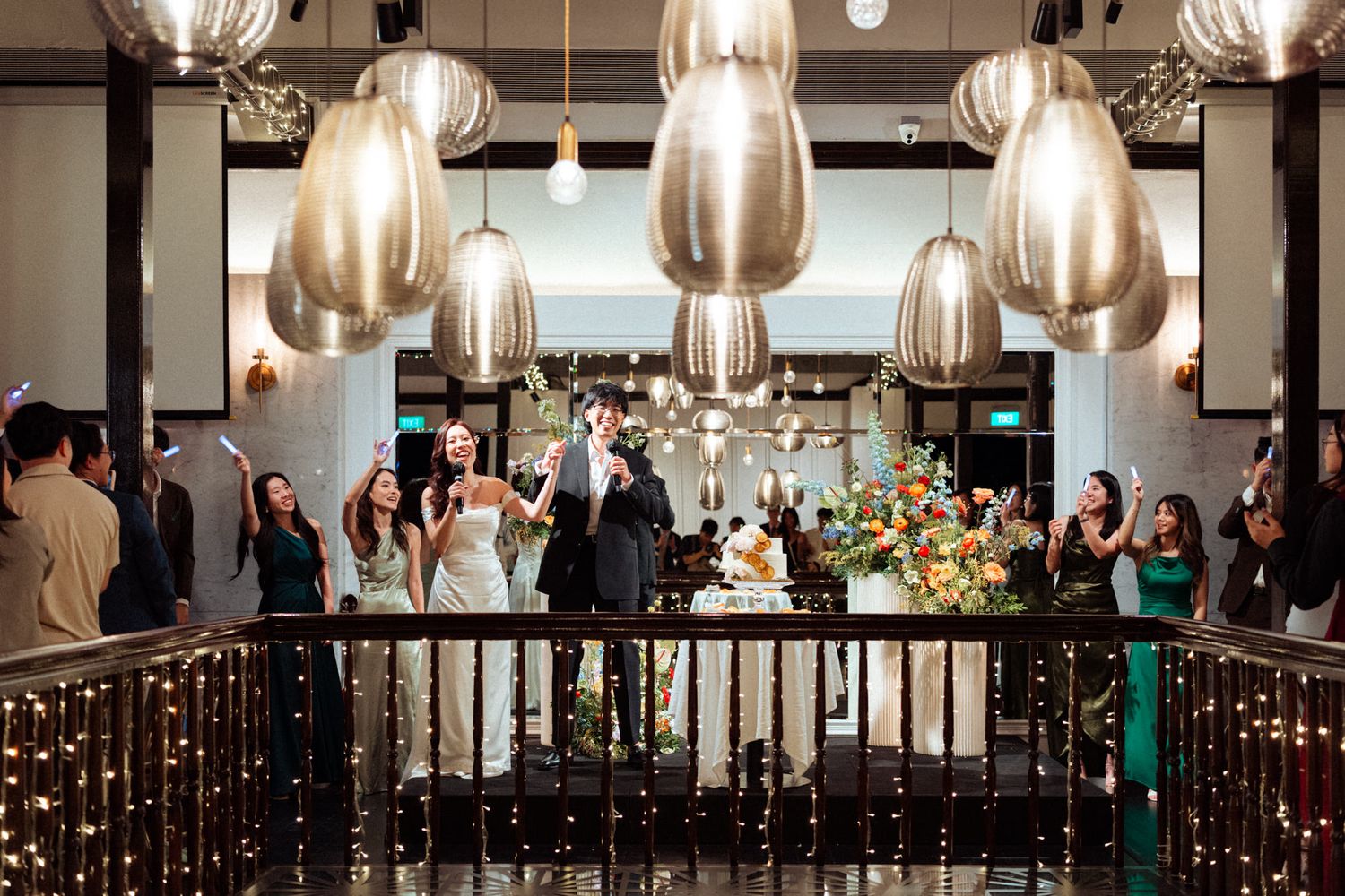 Elegant wedding reception with couple cutting cake under ornate pendant lights, surrounded by guests on a decorated balcony.