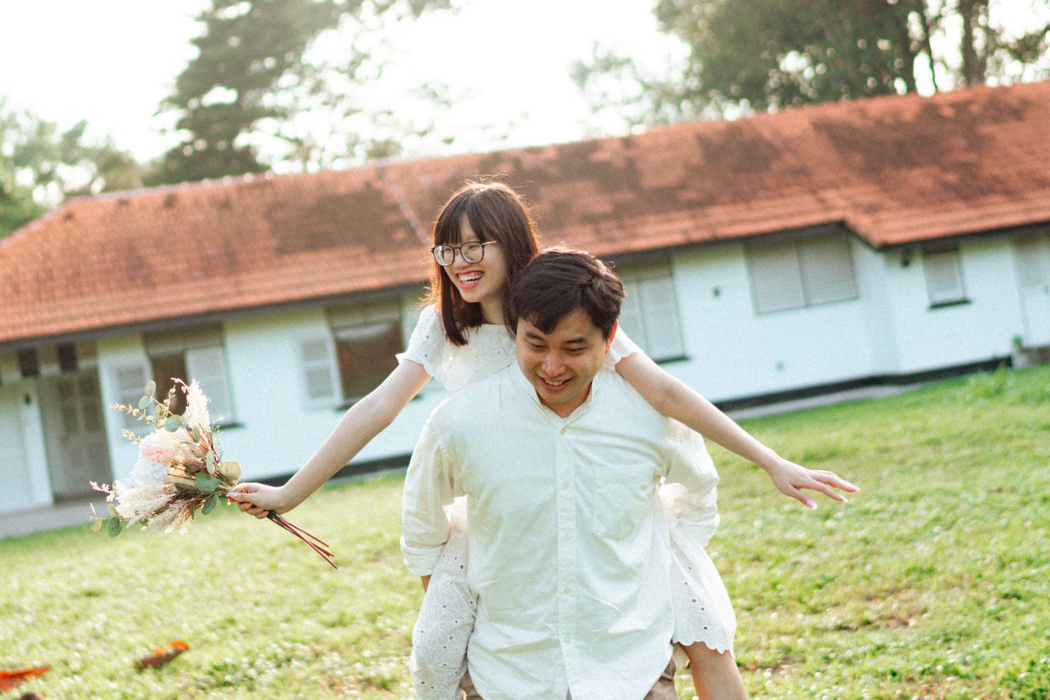 A couple in white outfits playfully runs and dances together on a grassy lawn in a romantic outdoor setting.