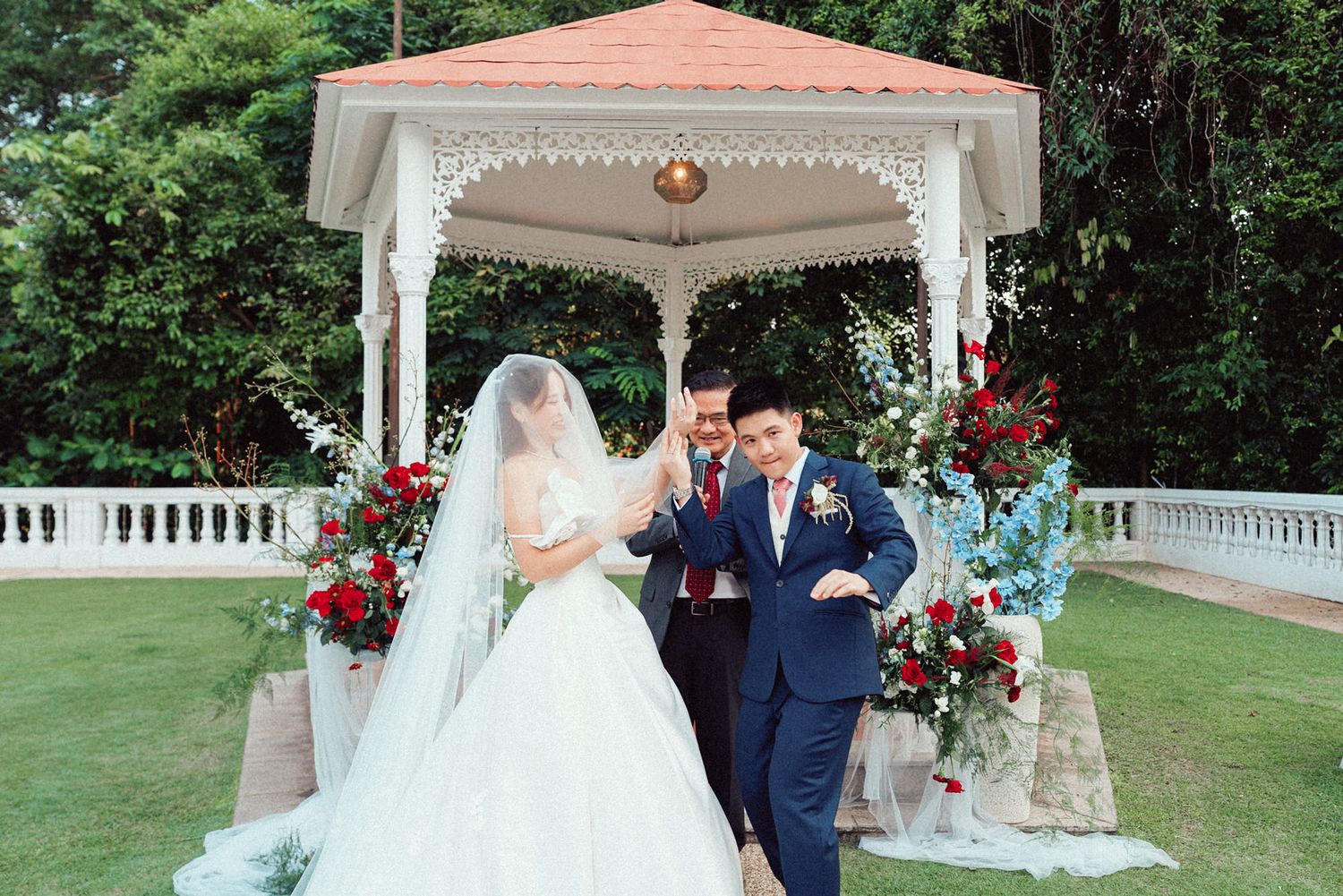 Elegant outdoor wedding ceremony with white gazebo, red floral arrangements, and lush green garden backdrop.