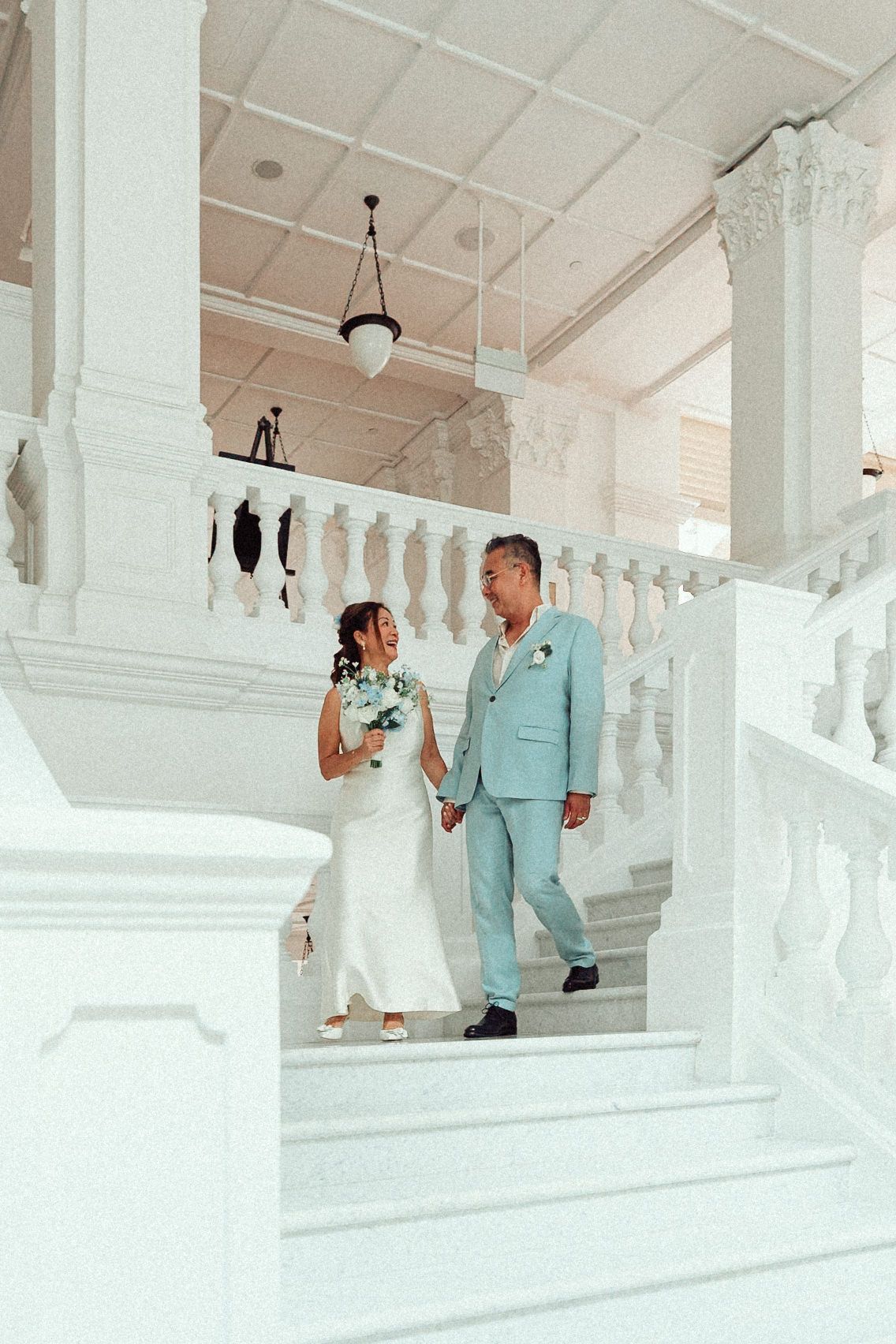 A couple poses playfully on white steps in formal attire during what appears to be a wedding photoshoot.