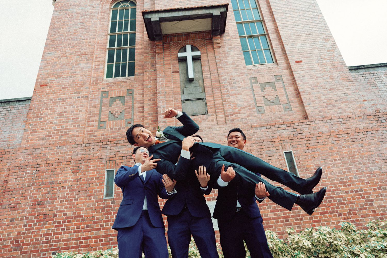 Groomsmen in navy suits playfully lifting someone in front of church.