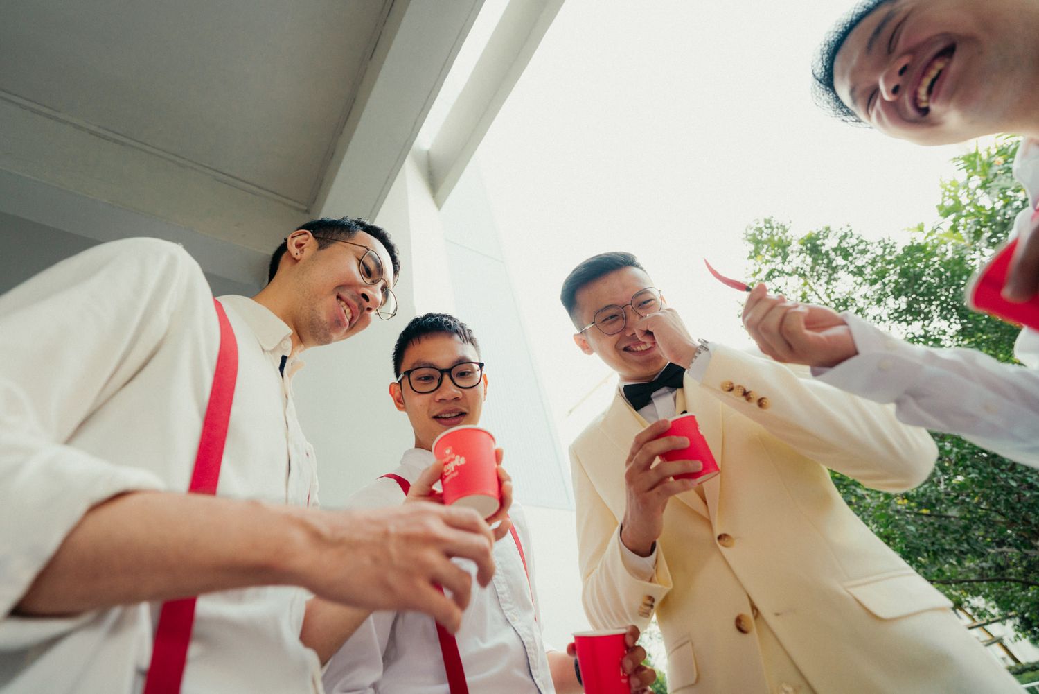 Four young men in formal attire holding red cups at an outdoor wedding celebration.