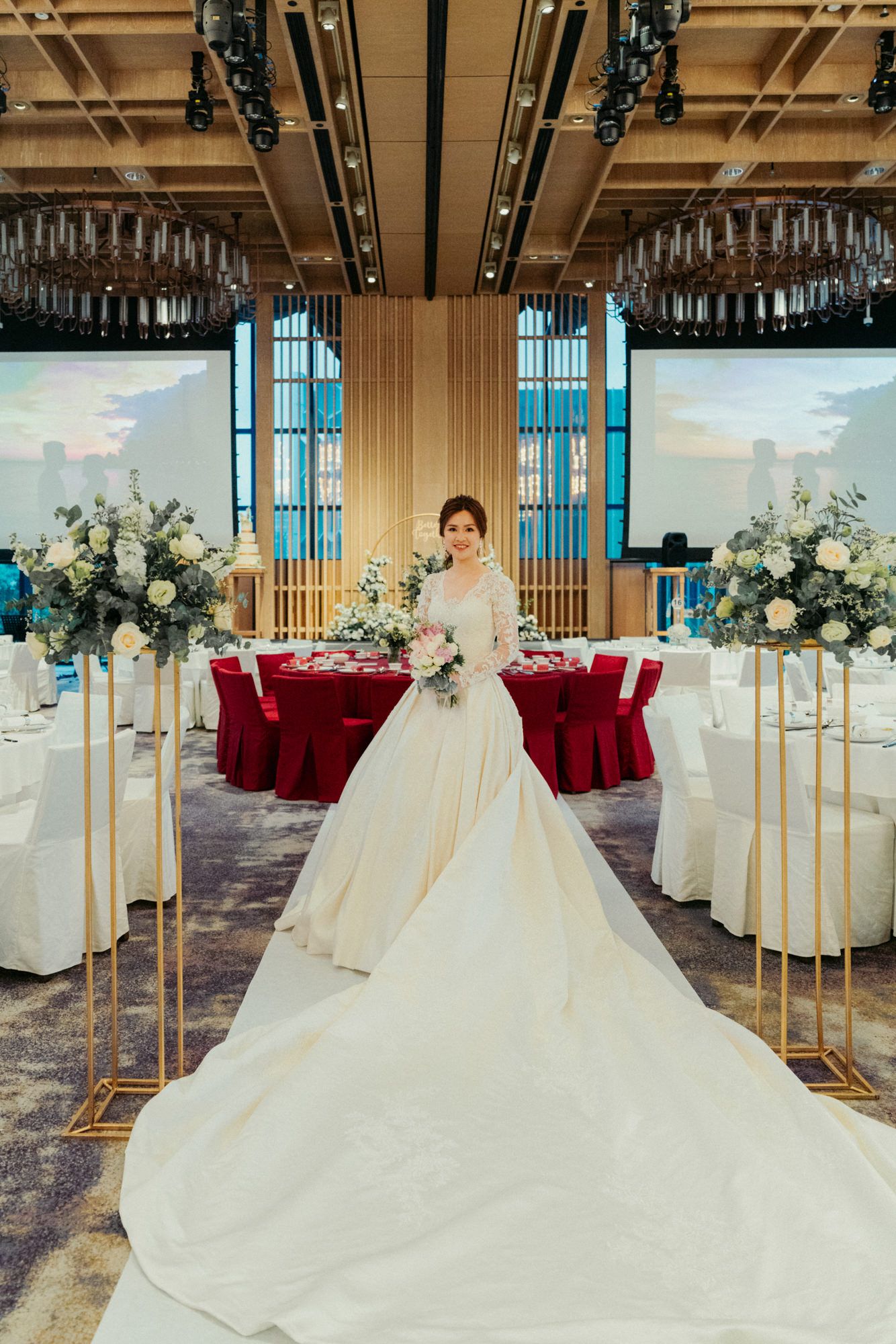 Bride in elegant white ball gown with long train stands between floral arrangements in a decorated wedding banquet hall.