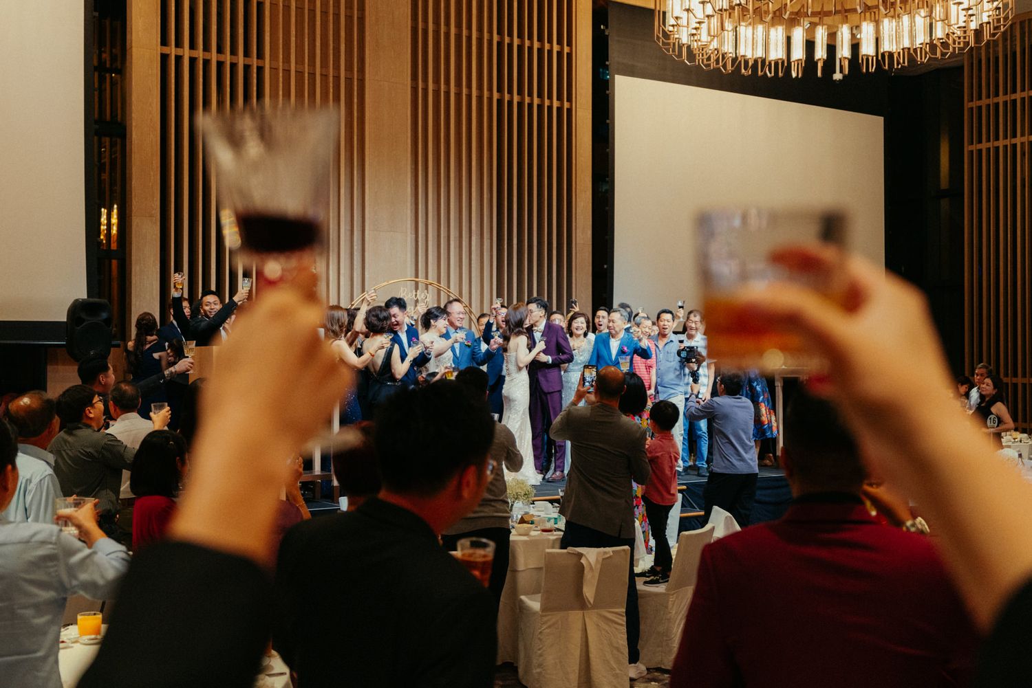 Guests raise glasses in a toast at an elegant wedding banquet hall with chandelier lighting.