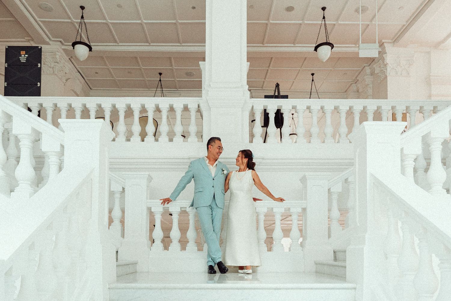 A couple descends an elegant white staircase with ornate balustrades in a grand indoor setting.