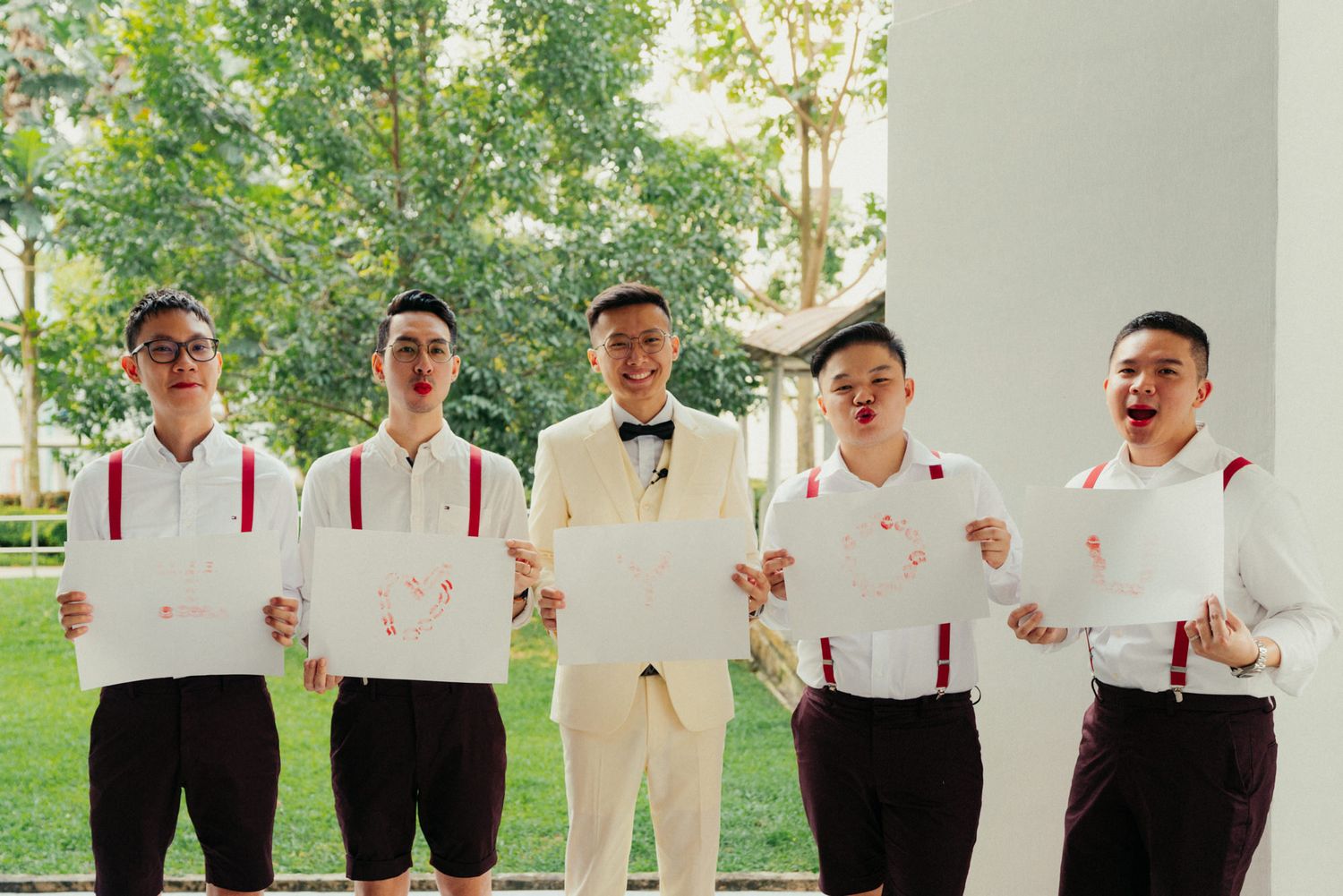 Groom in white tuxedo poses with groomsmen in white shirts and red suspenders holding signs outdoors.
