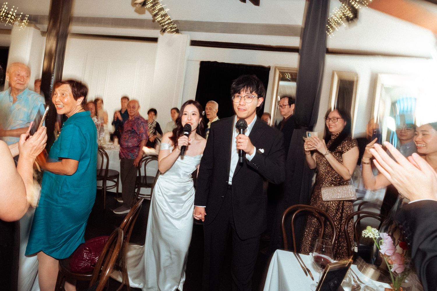 Couple singing with microphones at wedding banquet surrounded by applauding guests.
