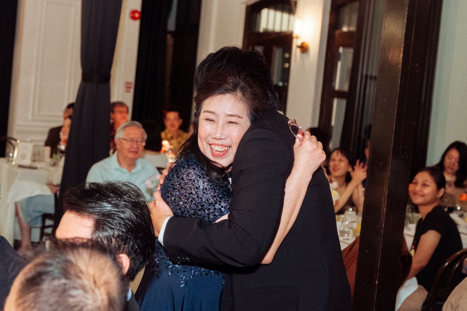 Two women share a warm hug at an elegant indoor event, surrounded by seated guests in a formal dining setting.