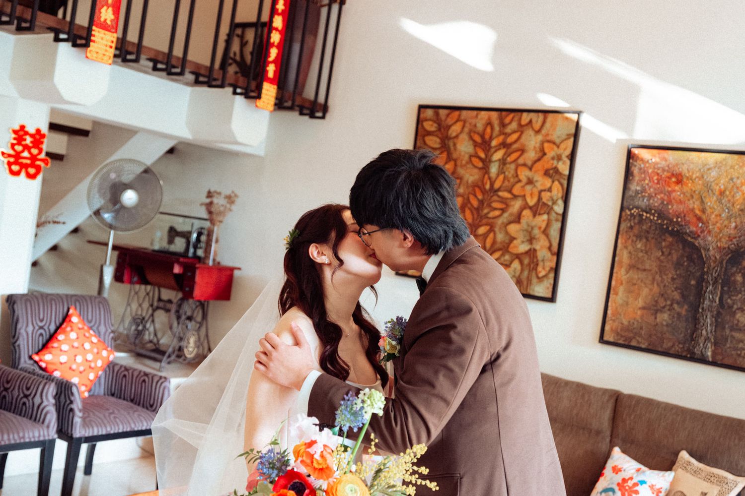 A couple shares a kiss in wedding attire, surrounded by colorful decor and artwork in an elegant indoor setting.