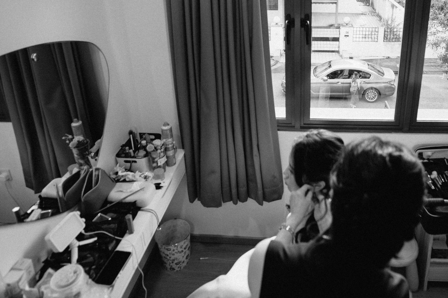 Black and white photo of a person doing makeup at a vanity mirror with curtains and a car visible through the window.