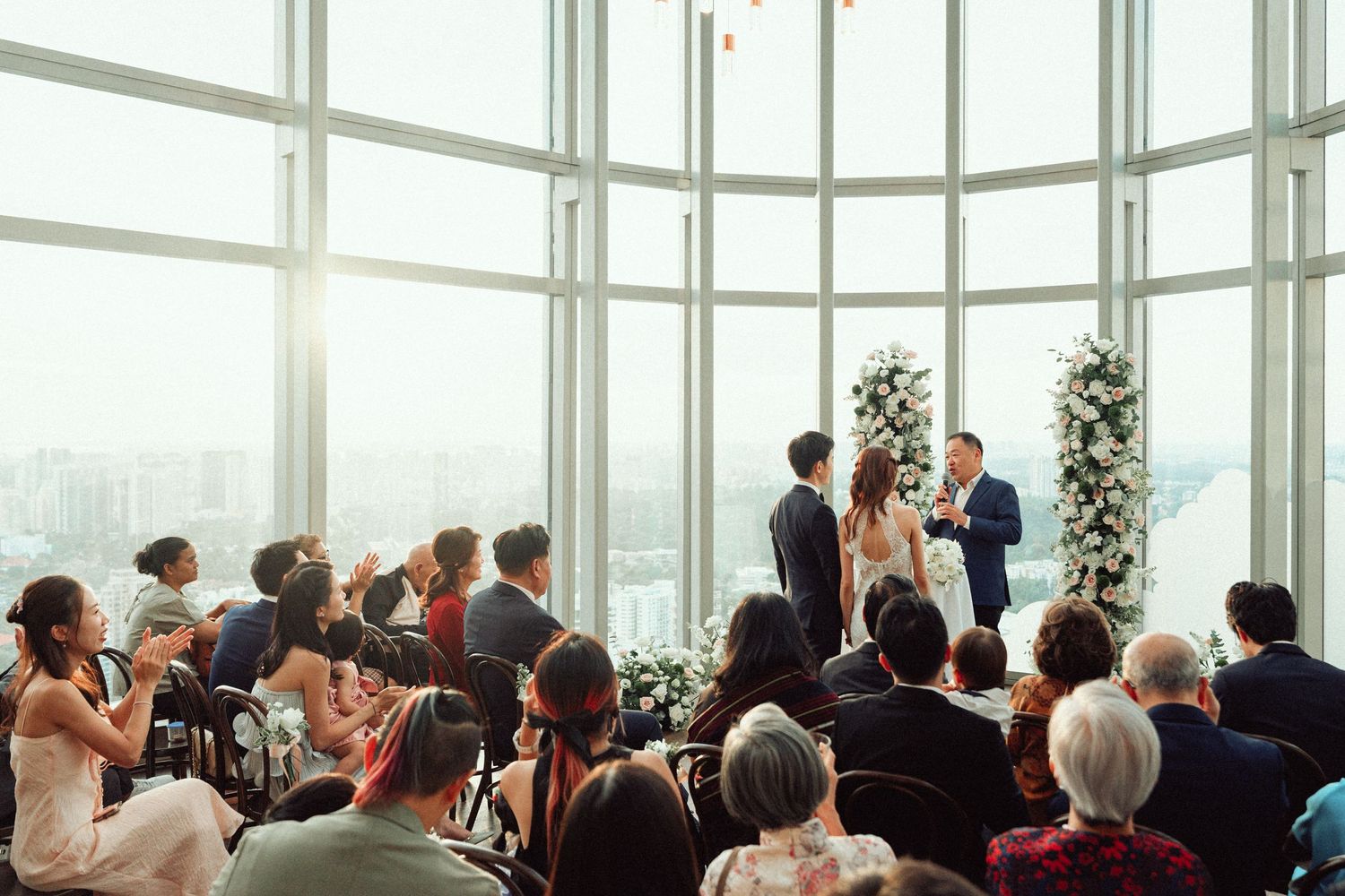 Wide shot of a wedding ceremony in a bright, glass-enclosed venue with city views.