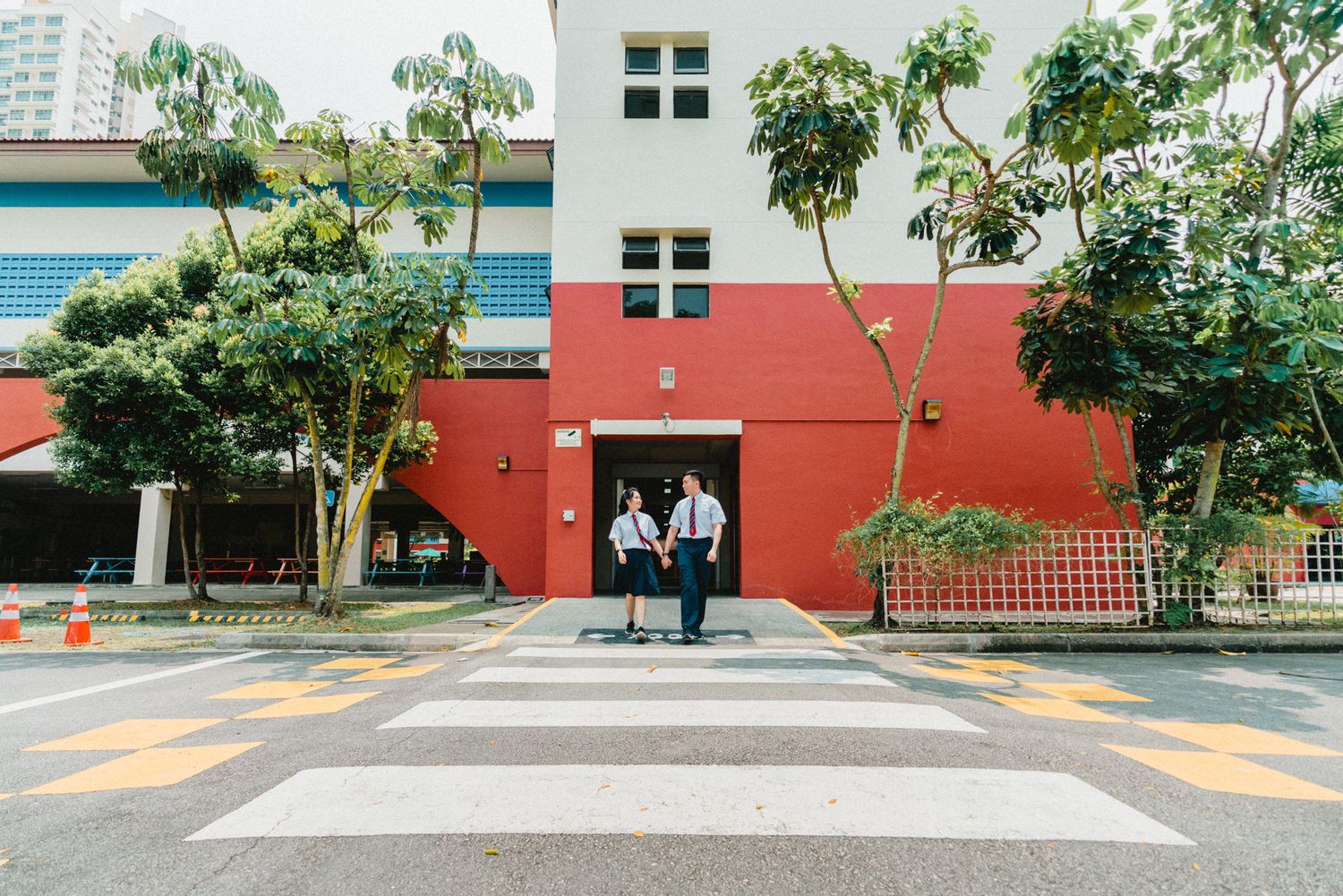 Students in uniforms standing in front of a red and white school building with trees.