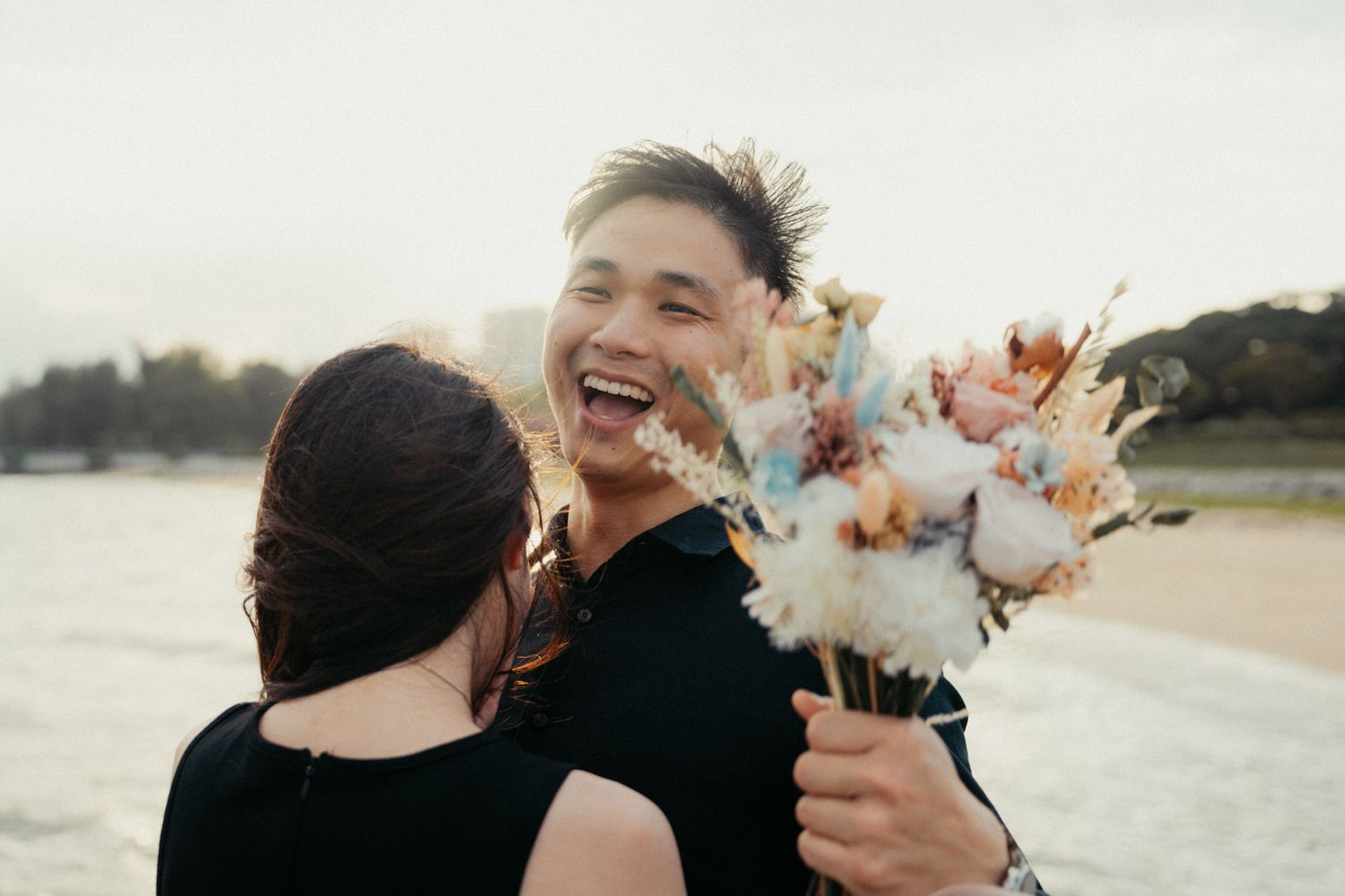 A couple laughs together while holding flowers on a scenic beach.