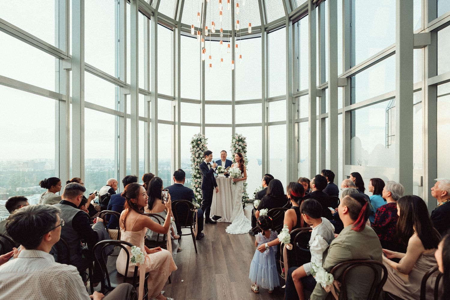 Elegant wedding ceremony in a glass-walled venue with city views, guests seated as couple exchanges vows at floral altar.