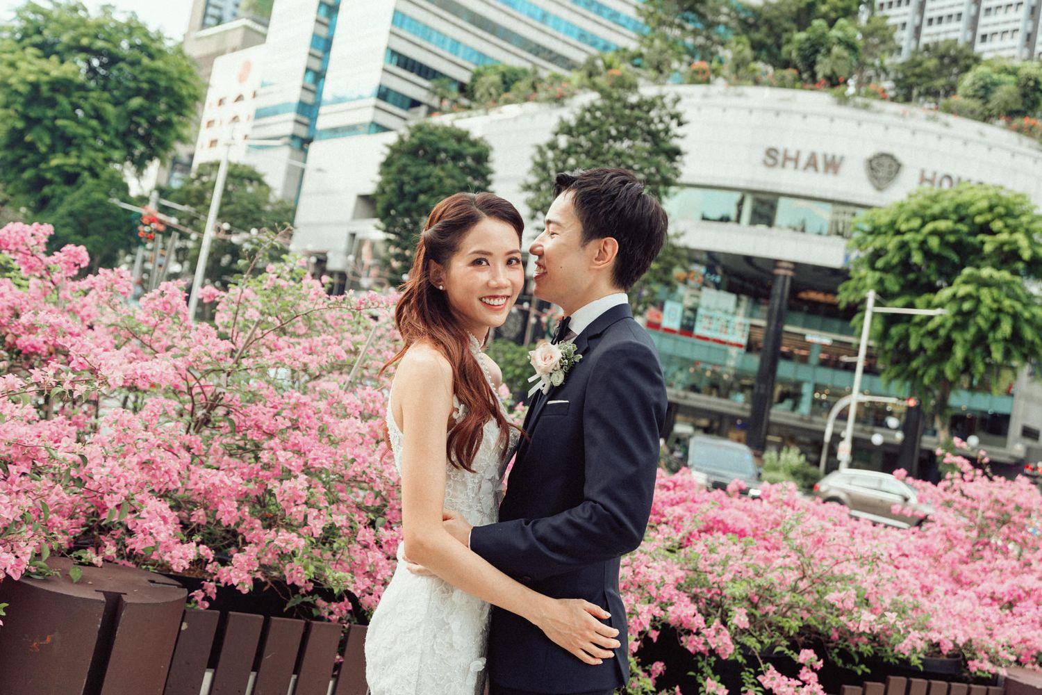 A couple in wedding attire embraces among pink flowers with urban buildings in the background.