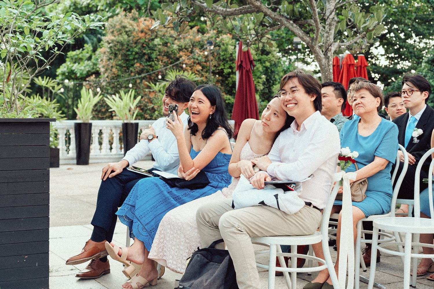 Wedding guests share laughter while seated in white chairs during an outdoor garden ceremony.