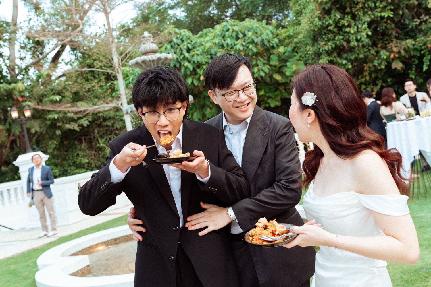 Three guests in formal attire share food and laugh together at an outdoor wedding reception on a green lawn.