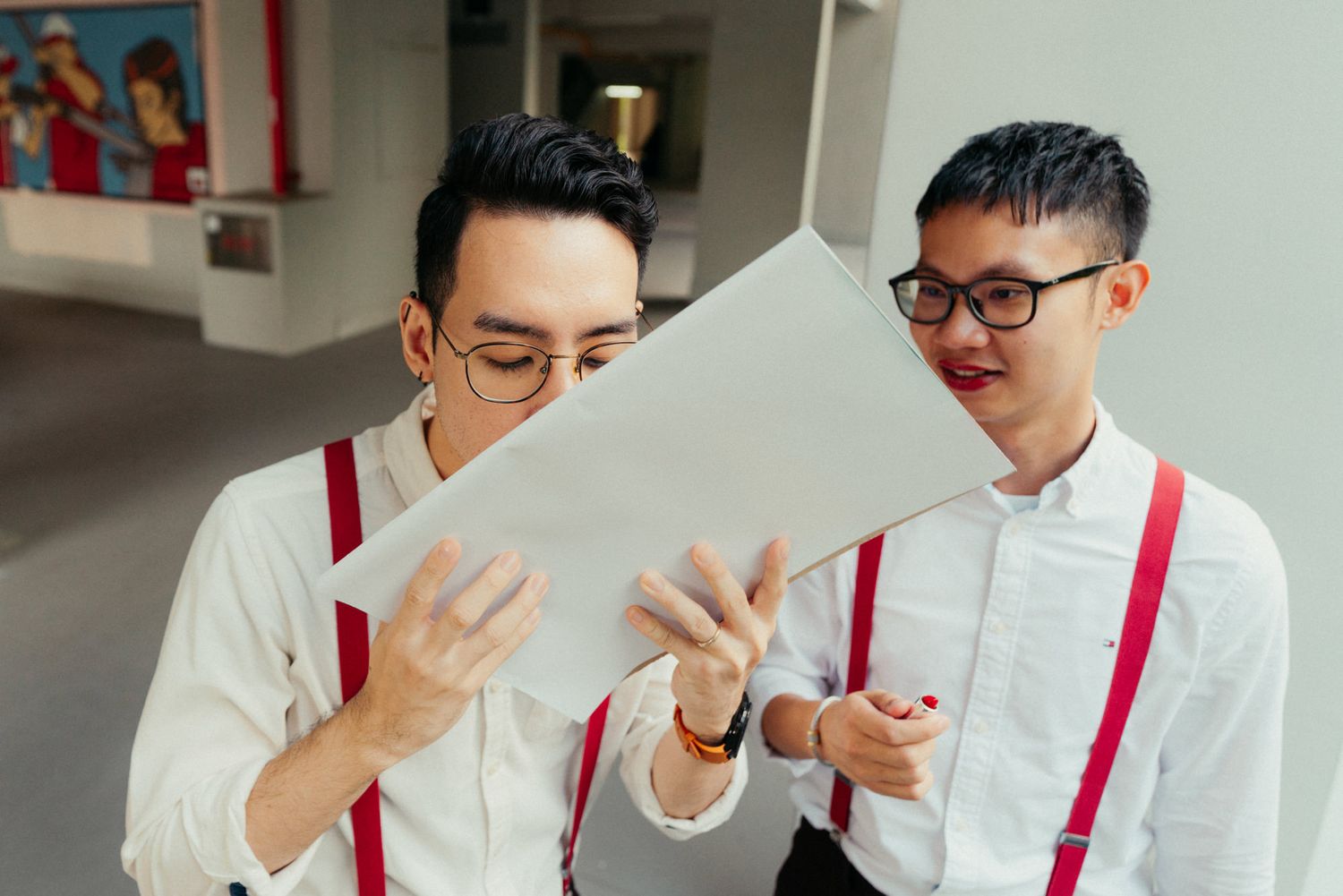Two young Asian men in white shirts with red suspenders reviewing documents together outdoors.