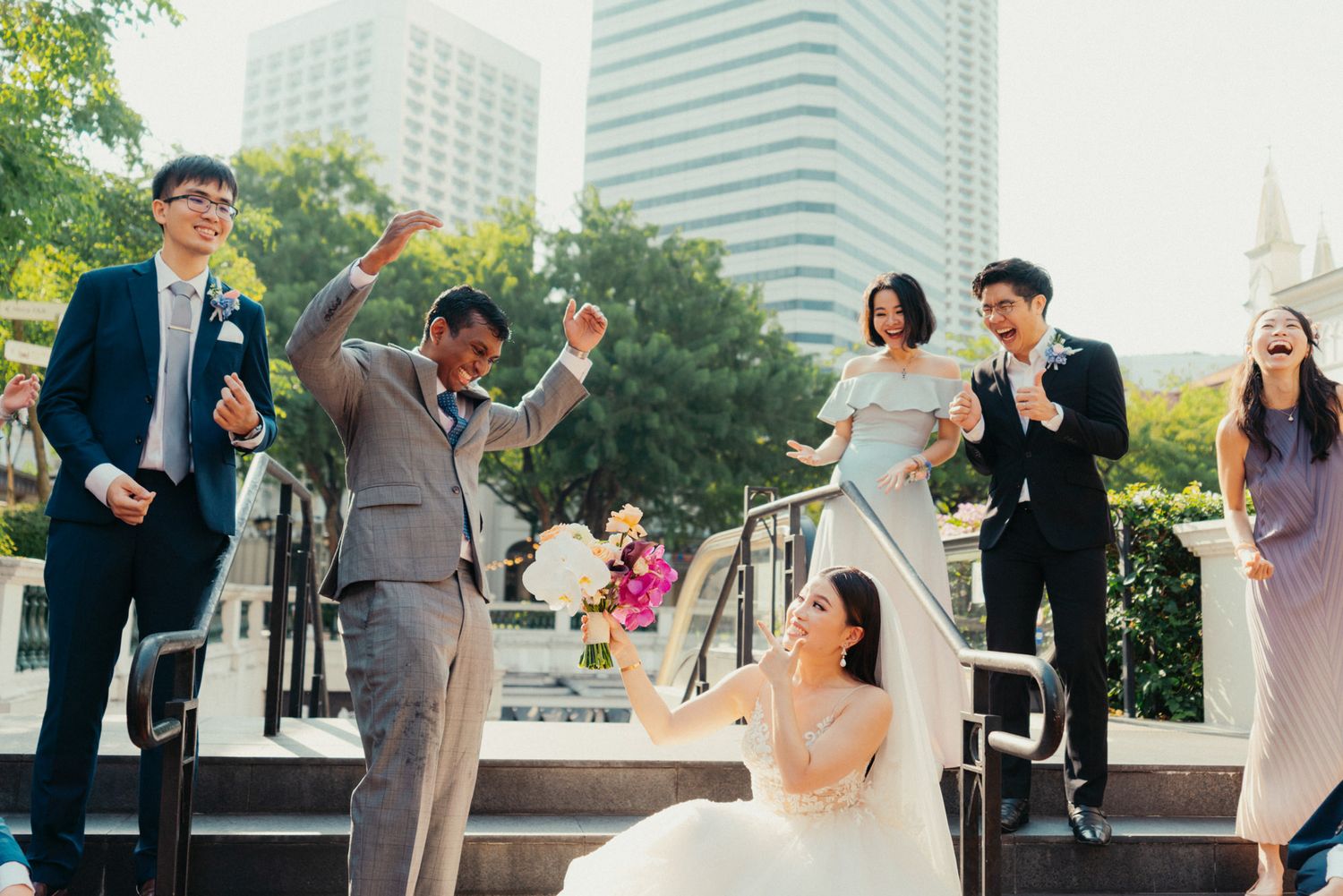 Joyful wedding party celebrating on outdoor steps with bride holding bouquet, groom raising arms, and guests cheering.