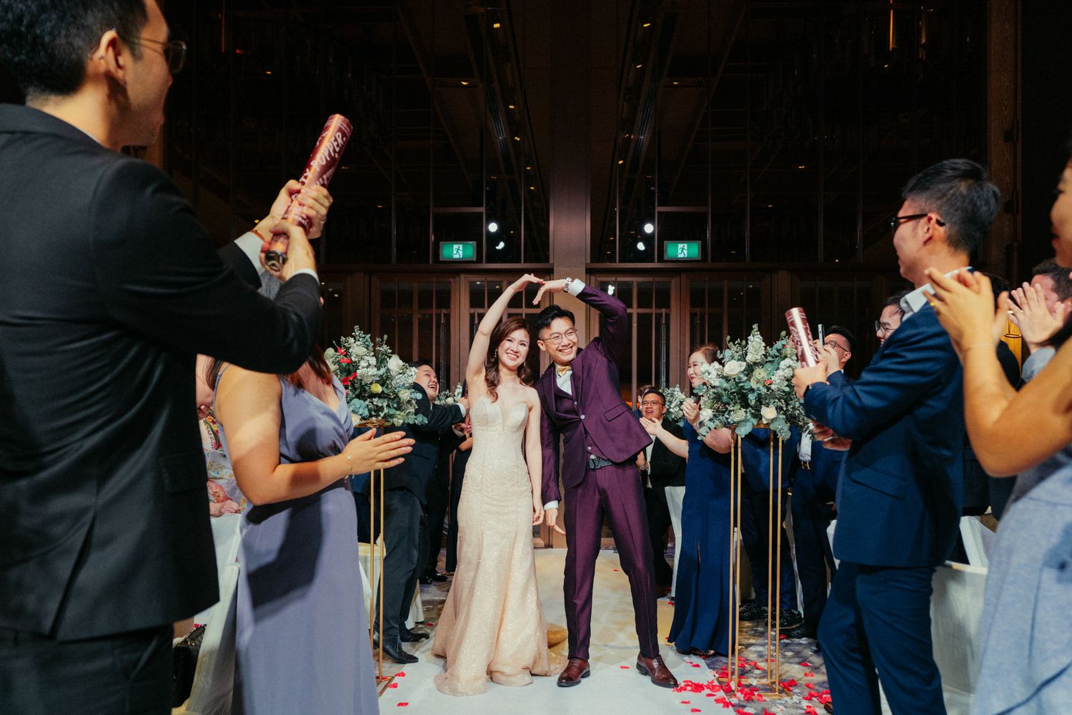 Bride and groom celebrate their wedding reception exit, with guests cheering and tossing petals in an elegant venue.