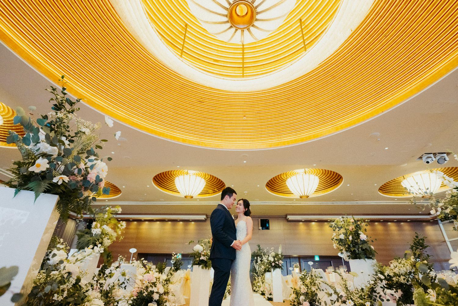 Elegant wedding couple embracing under golden domed ceiling with floral arrangements in a luxurious ballroom.