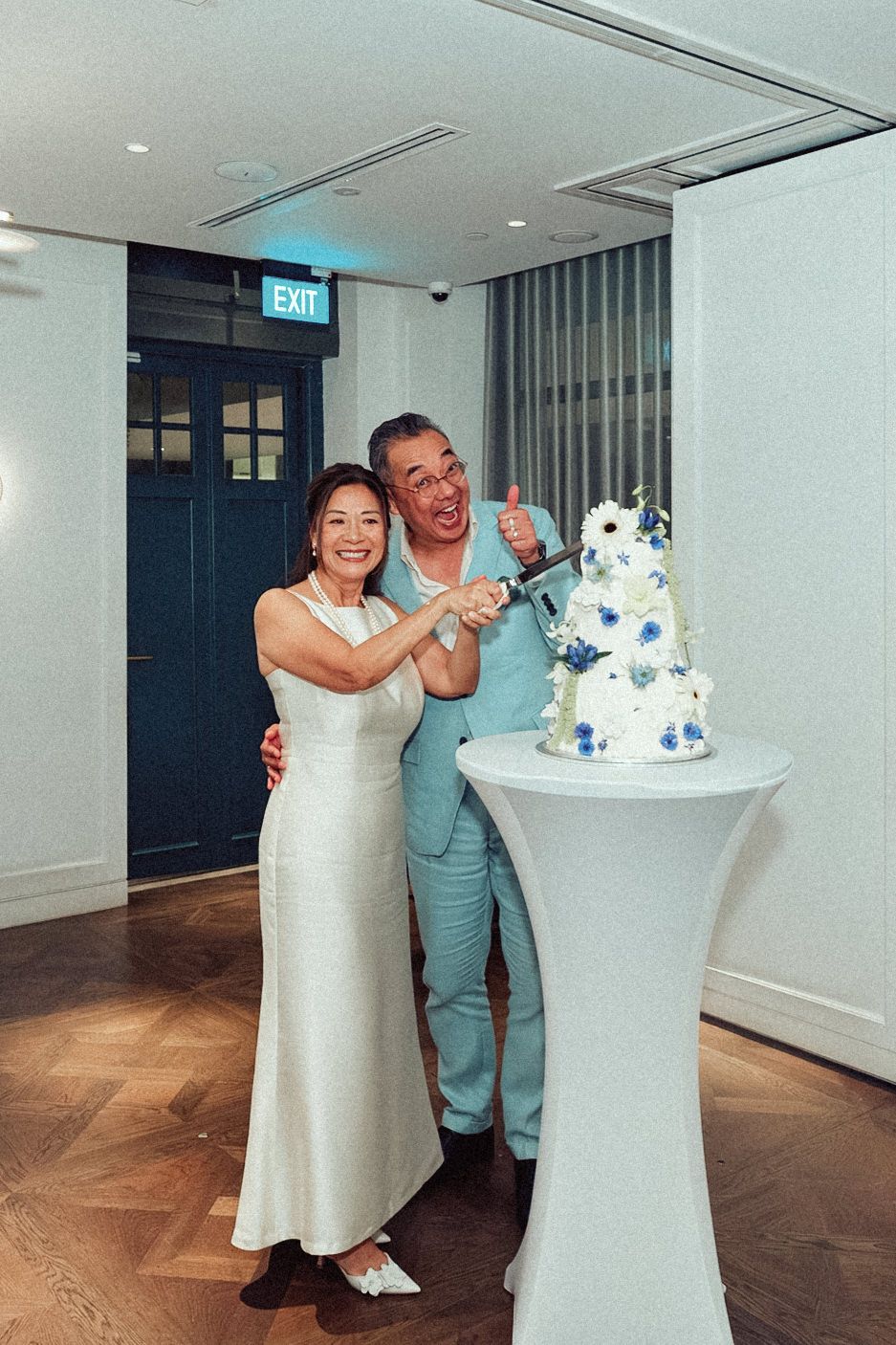 A couple in wedding attire playfully poses and dances next to their elegant white and blue wedding cake.