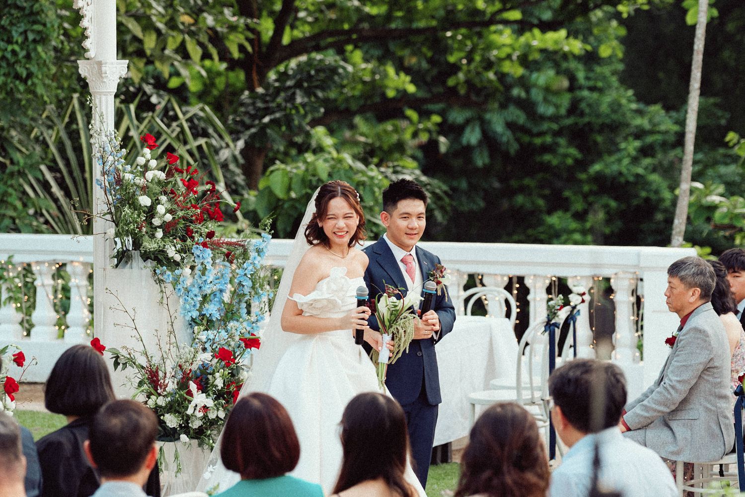 Wedding ceremony takes place under a white gazebo decorated with red roses in an outdoor garden setting.