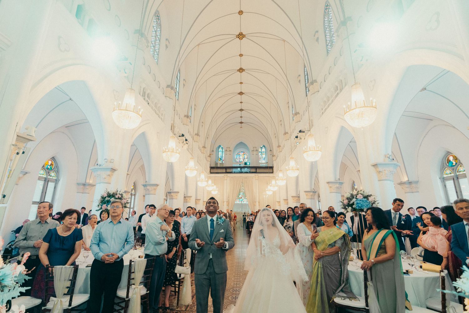 Bride and groom walk down the aisle of a stunning white cathedral church surrounded by wedding guests.