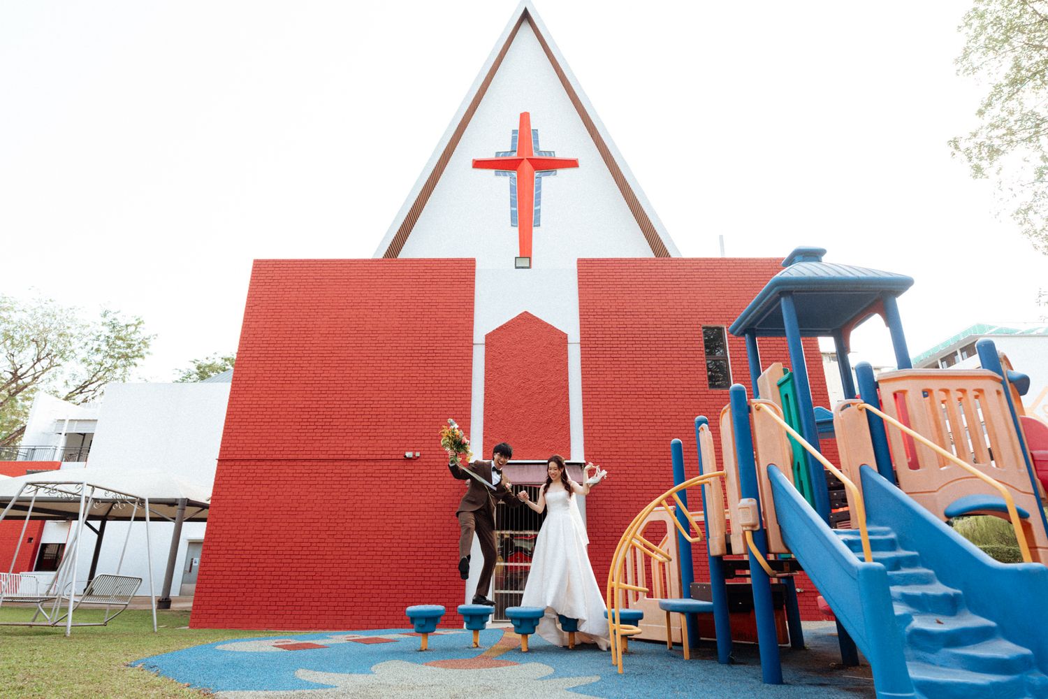 Couple celebrating in front of a red and white church with a cross, near a colorful playground on a sunny day.