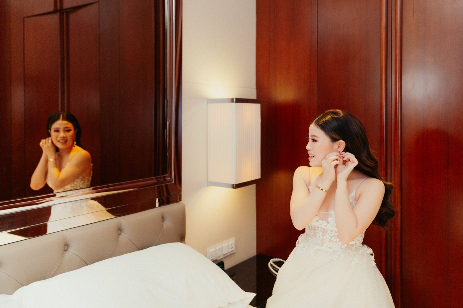 Bride in white wedding gown putting on earrings while seated before a mirror in an elegant hotel room.