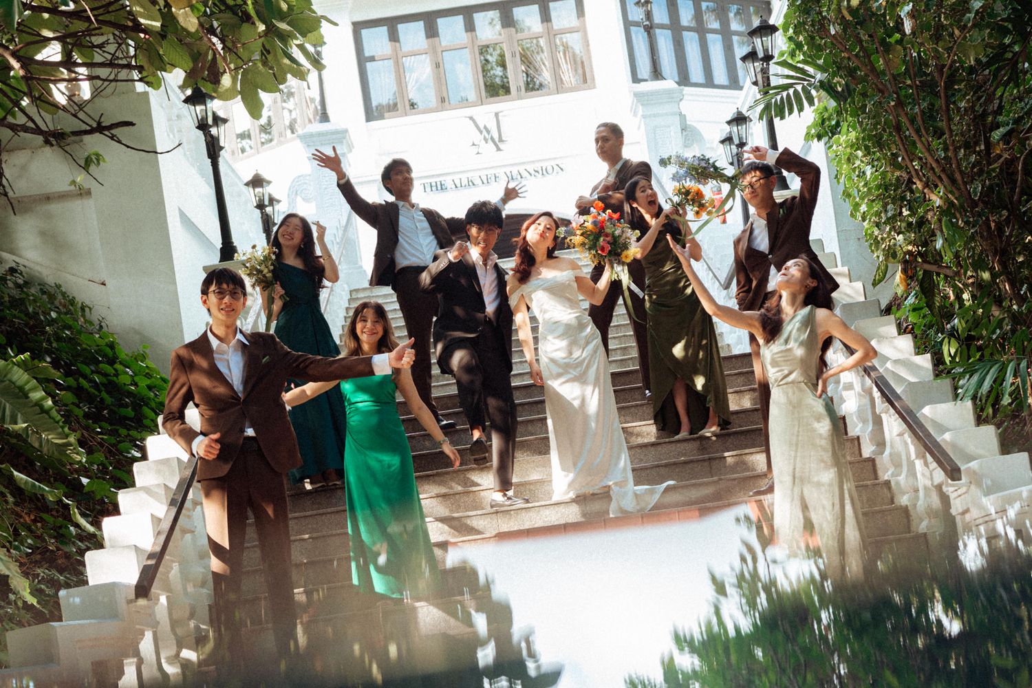 Joyful wedding party celebrating on white steps outdoors, bride in white gown surrounded by cheering guests in formal attire.