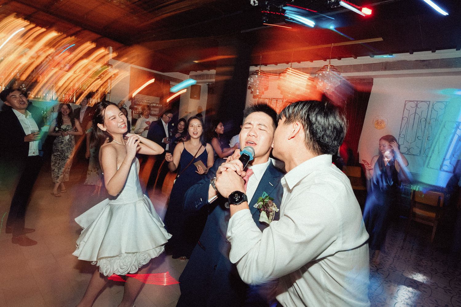 Lively scene at a party venue with people singing karaoke under colorful lighting in a rustic wooden ceiling space.