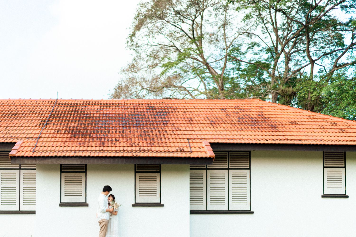 Single-story building with red tile roof and white walls set against trees in a sunny outdoor setting.