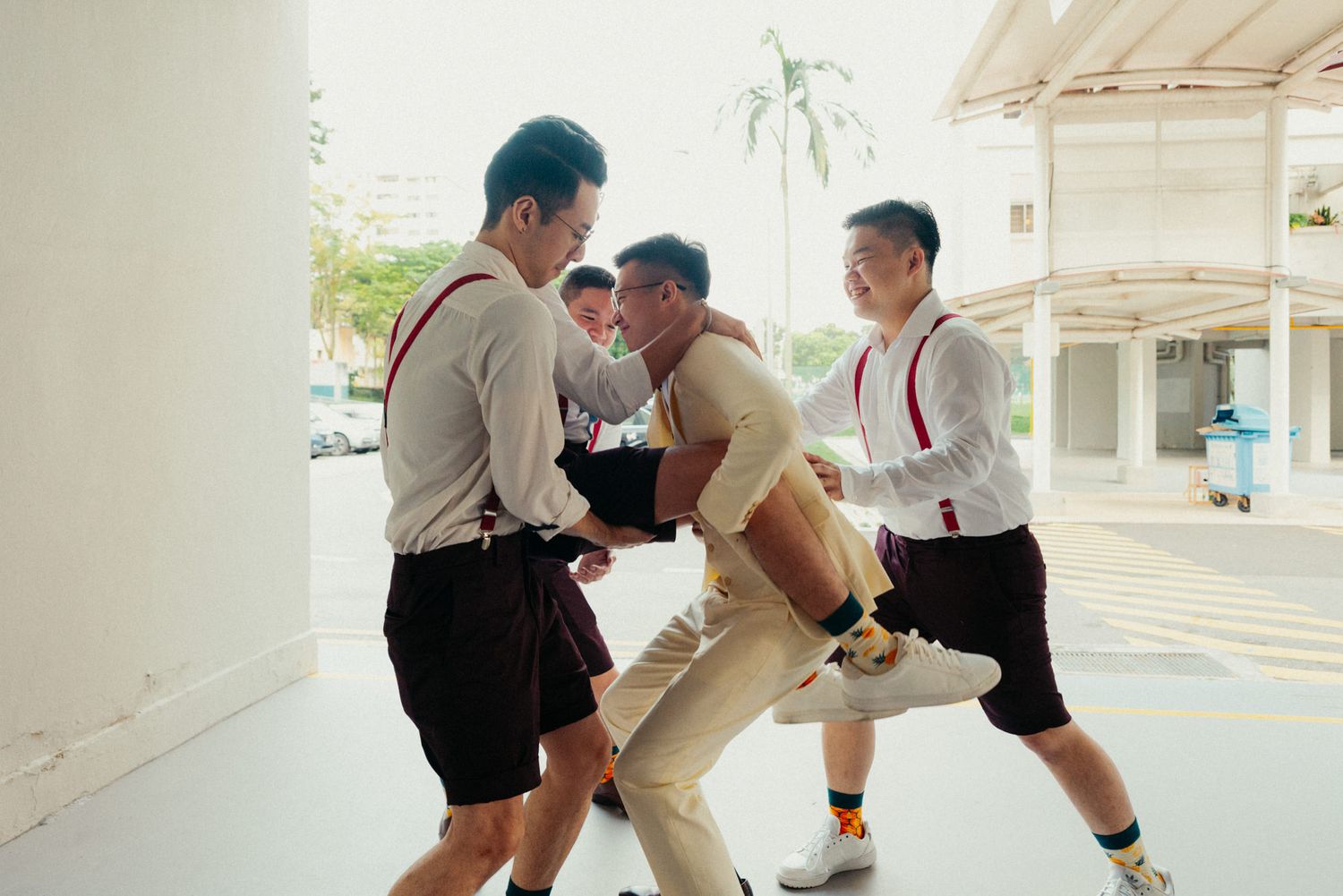Students in white shirts and suspenders playfully wrestling outdoors near a building in a school setting.
