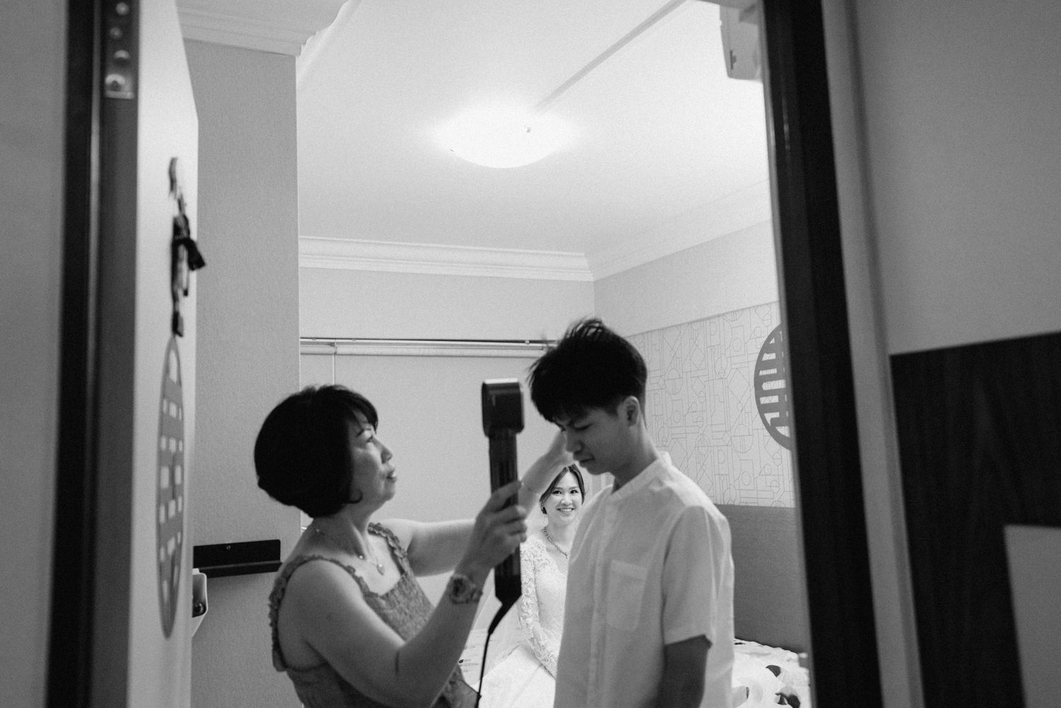 Black and white photo of a woman adjusting a young man's collar in a bright room, captured through a doorway.