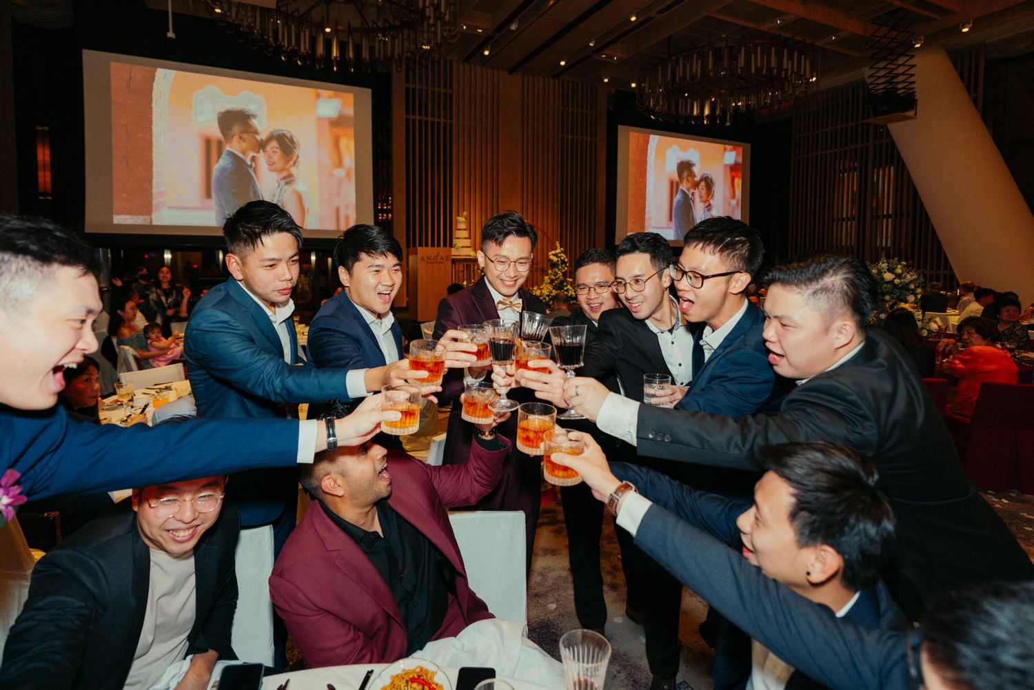 Group of men in suits toasting with beer glasses at a wedding reception banquet in a decorated hall.